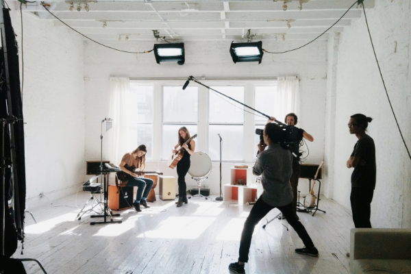 A music band is performing in a bright, white studio with large windows. A person is operating a boom microphone while another films with a camera. The band members, playing instruments including guitar and drums, create an electrifying atmosphere as the scene captures the essence of a small business in Melbourne. | Ven Agency